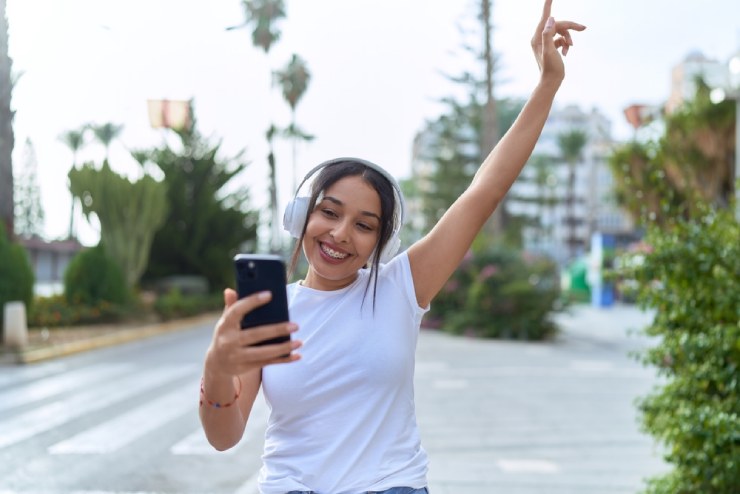 Young woman wearing braces smiles while listening to music via white over-the-ear headphones while walking down the street in California