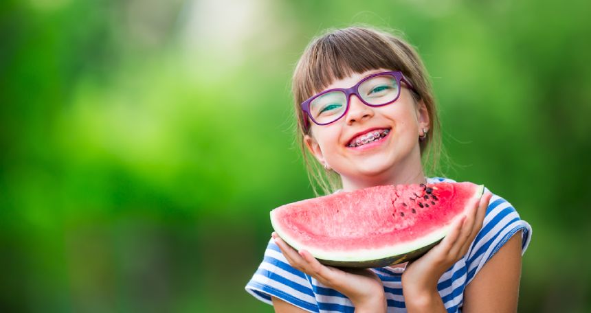 A child wearing glasses and braces smiles while holding a large wedge of watermelon.