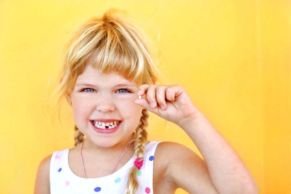 Girl on Yellow Background Holding Missing Bottom Front Tooth and Smiling