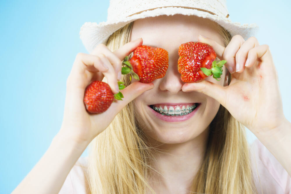 A young woman with braces in a hat playfully holds strawberries up to her eyes against a sky-blue background
