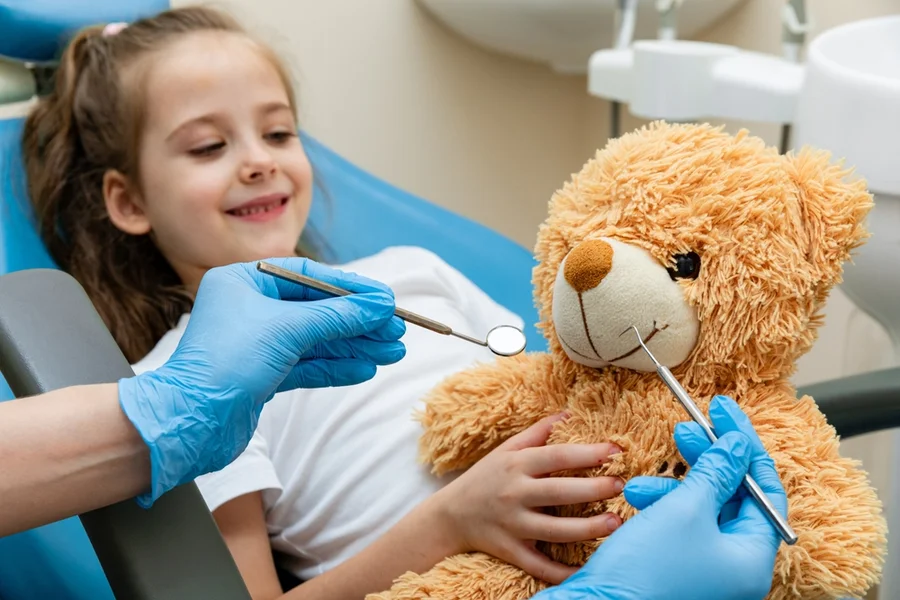 Dentist playfully examines a teddy bear’s mouth with instruments to make a young girl feel comfortable