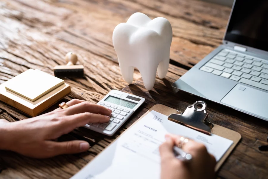 Giant tooth placed next to a rubber stamp, sticky note pad, calculator, laptop, and clipboard with a person’s hand filling out a dental insurance form