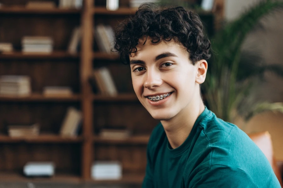 A teenage boy in a teal shirt wearing braces, sitting in a library, smiles