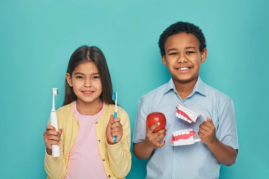 Young girl holds electric and manual toothbrush next to young boy holding a model of teeth and an apple