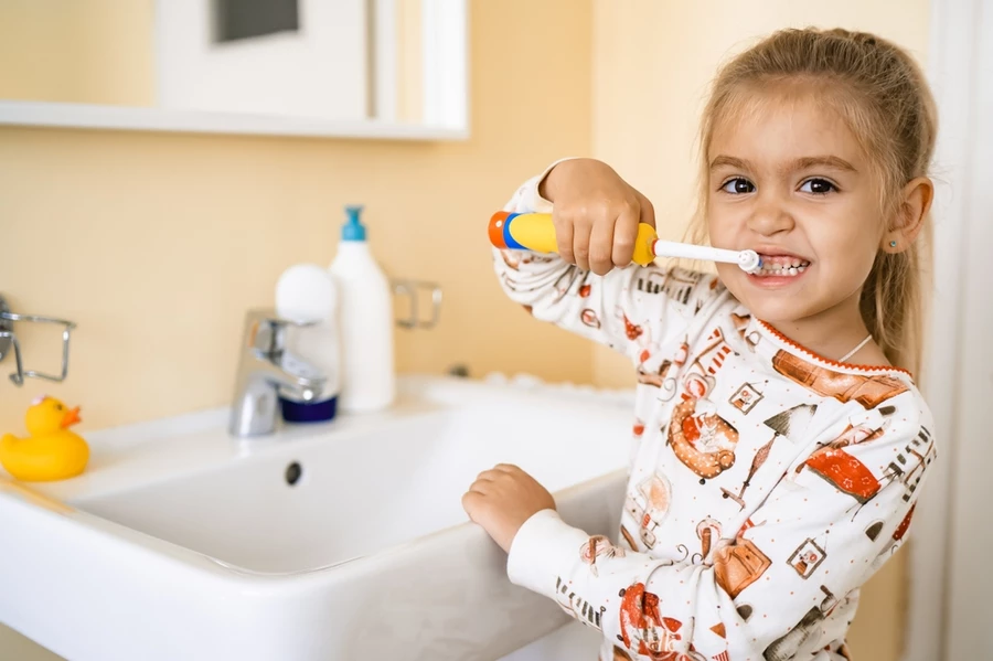 Young girl in PJs uses colorful yellow blue and orange electric toothbrush for kids