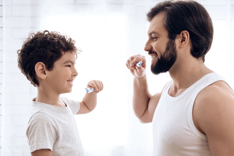 Father and son look at each other smiling while brushing their teeth