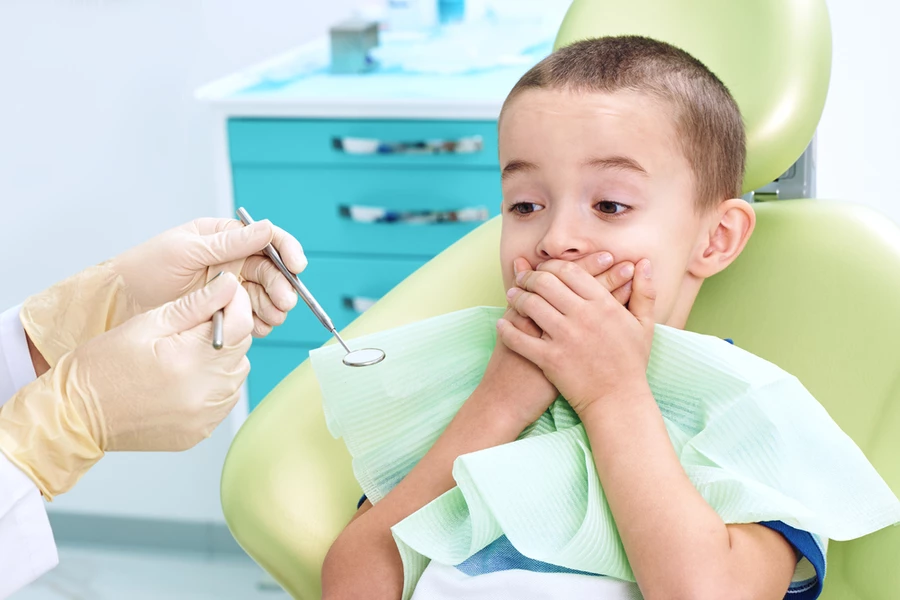 Young boy sits in dental chair and covers mouth in fear