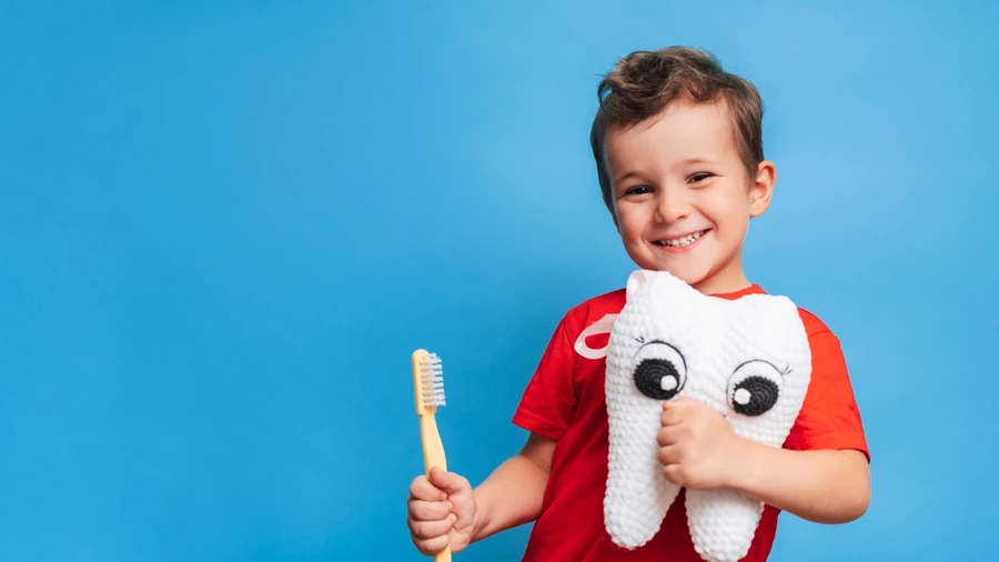 Young boy wearing a red shirt holds a toothbrush in one hand and a plush tooth in another while smiling