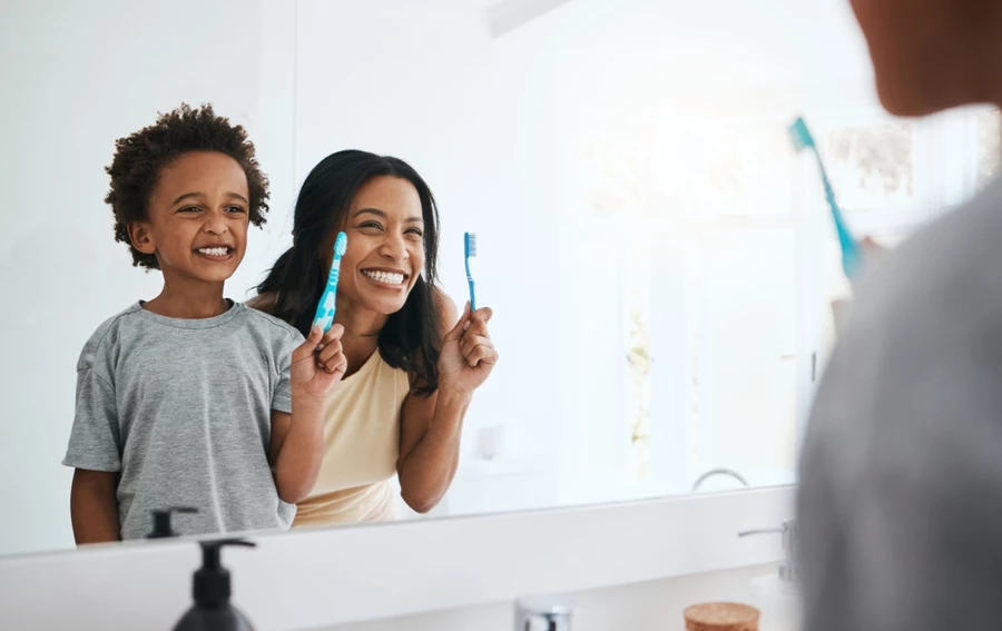 Young boy smiles holding a toothbrush next to his mom smiling demonstrating the importance of dental hygiene for kids